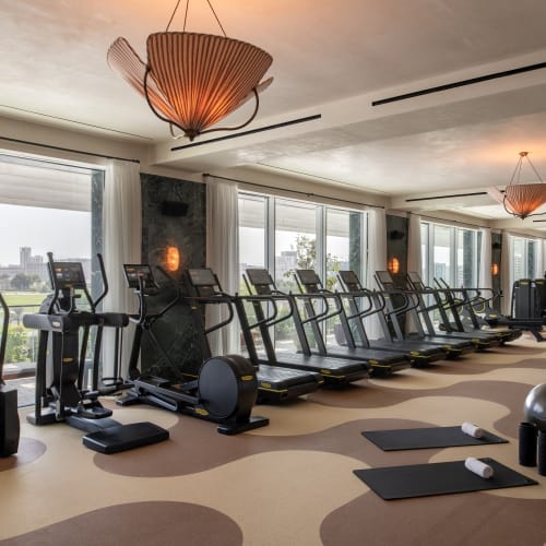 Ein geräumiges Fitnessstudio im The Ned Doha mit mehreren Laufbändern und anderen Trainingsgeräten, die neben großen Fenstern mit Stadtblick in Reihen angeordnet sind.