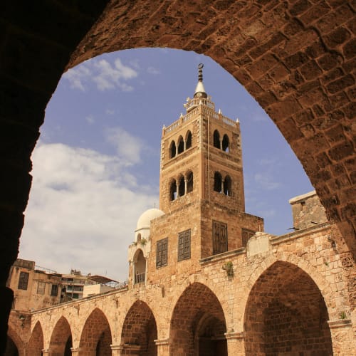 A historic stone tower viewed through an arched brick structure in Tripoli, Lebanon.