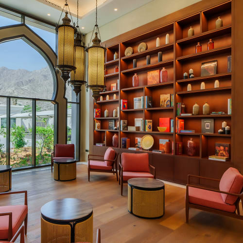 Ein gemütlicher Lounge-Bereich in den Naäma Beach Villas und Spa mit hölzernen Regalen voller Dekorationsgegenstände, korallfarbenen Polstersesseln, runden Holztischen und kunstvollen hängenden Laternen sowie einem großen Fenster mit Blick auf eine Berglandschaft.