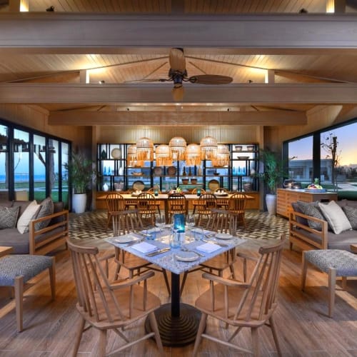 Elegant restaurant with wooden furniture and large windows overlooking the beach at sunset.