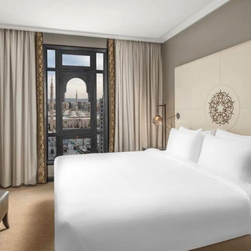 A hotel room with a large bed, a window with curtains, and a view of a mosque dome and cityscape in the distance.