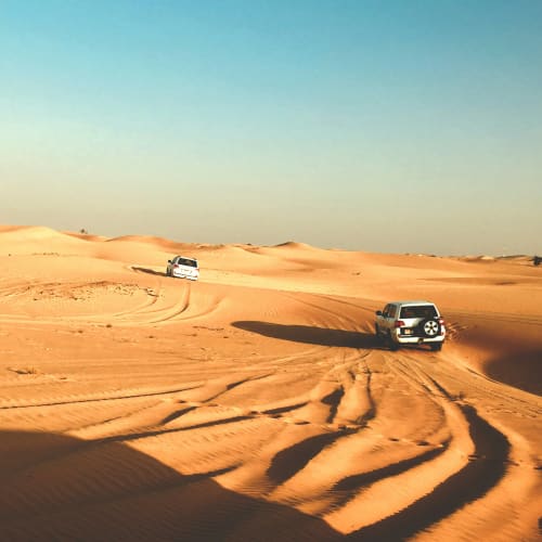 Zwei weiße SUVs fahren über die Sanddünen in der Wüste von Abu Dhabi mit sichtbaren Reifenabriebspuren.