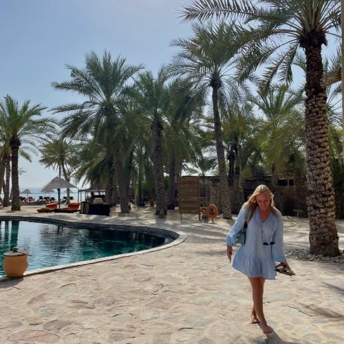 A woman walking along a stone pathway near a pool, surrounded by tall palm trees under a clear blue sky.