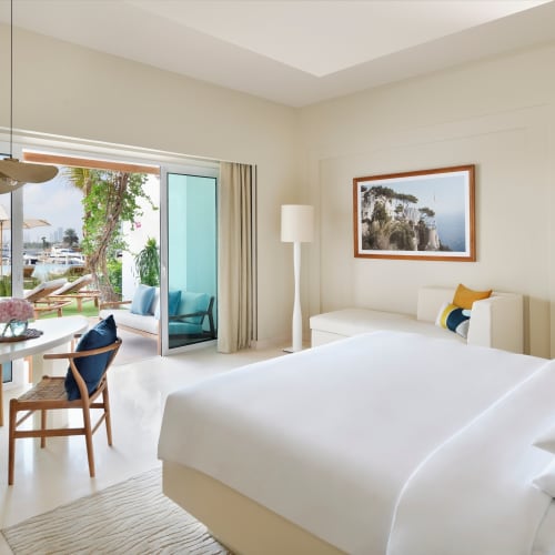 Helles und geräumiges Hotelzimmer mit großem Bett, Sitzbereich und Tisch mit Blick auf eine Terrasse mit Liegestühlen und Wasserblick umgeben von Grün.