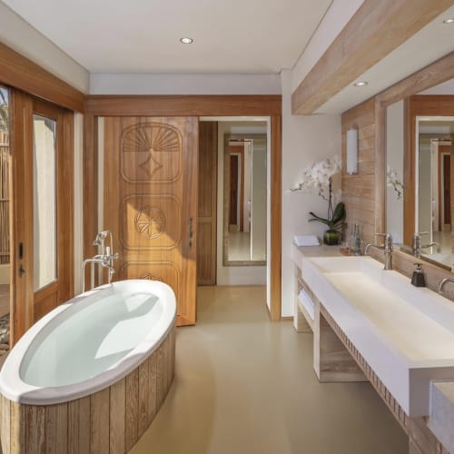 Ein modernes Badezimmer mit einer Badewanne mit Holzrahmen, die mit Wasser gefüllt ist, großen Spiegeln, zwei Waschbecken und einer Tür, die zu einem Außenbereich mit Bambuszaun führt.