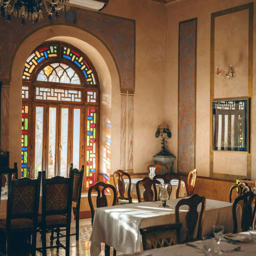 Elegantes Speisezimmer mit kunstvollem Buntglas-Tür, die Sonnenlicht hineinlässt, gedeckte Tische und antike Stühle.
