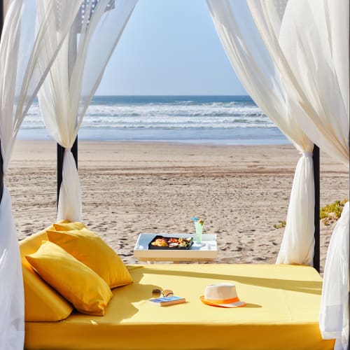 A beach cabana with a yellow mattress and pillows, white curtains tied to black posts, overlooking a sandy beach and ocean waves under a clear blue sky.