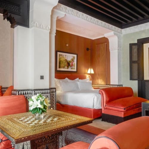 Ein luxuriöses Hotelzimmer mit einem dekorierten Bett, orangefarbenen Sofas, einem Holztisch mit Blumenarrangement und marokkanischem Dekor.