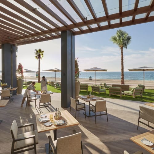Außenbereich eines Strandrestaurants mit Palmen, Tischen und Stühlen, Blick auf das Meer unter einer teilweise schattigen Überdachung.