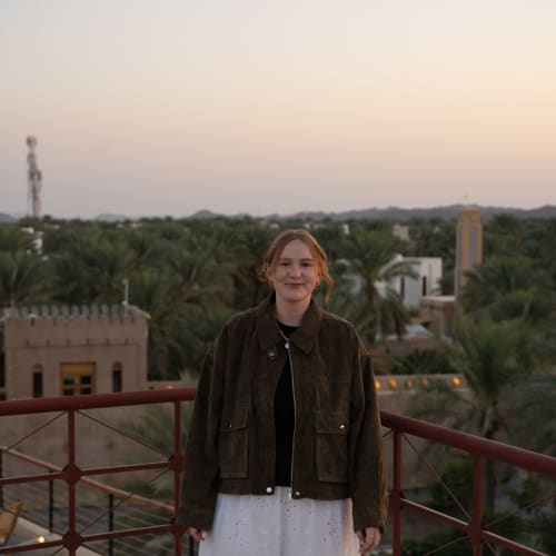 Eine Frau steht auf einem Balkon mit roten Geländern und blickt bei Sonnenuntergang auf eine grüne Landschaft mit Palmen und traditionellen Gebäuden.
