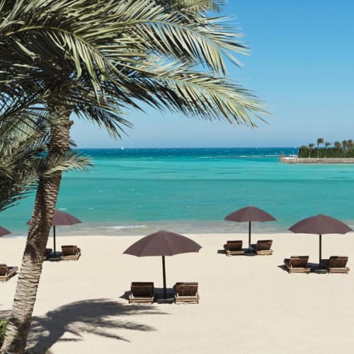 Eine tropische Strandlandschaft mit Palmen, mehreren braunen Sonnenschirmen und Liegestühlen auf weißem Sand, mit Blick auf türkisfarbenes Wasser und einen klaren blauen Himmel.