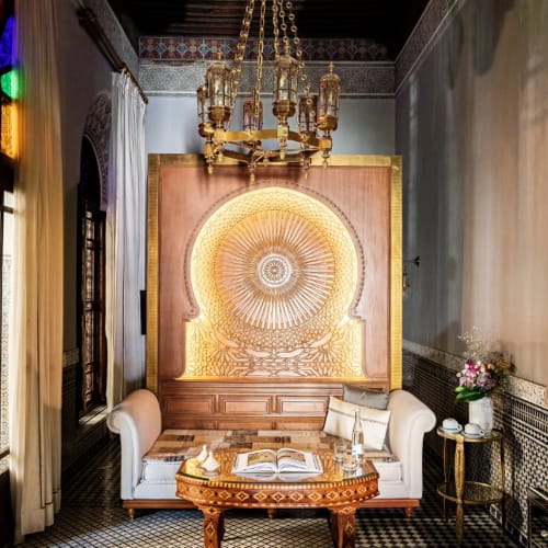 Luxurious lounge with a decorative golden wall feature, a chandelier, and elegant furniture including a sofa, table, and side table with flowers.