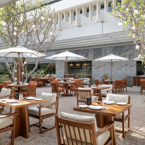 Außenbereich mit Esstischen und Stühlen aus Holz mit Polsterung, großen Sonnenschirmen und Bäumen, die Schatten in einem städtischen Innenhof spenden.
