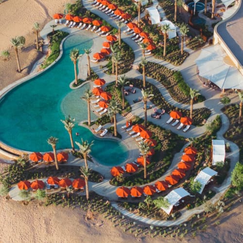 Eine Luftaufnahme eines Resorts mit großem Swimmingpool, umgeben von orangefarbenen Sonnenschirmen und Liegestühlen, mit Palmen und Wegen.