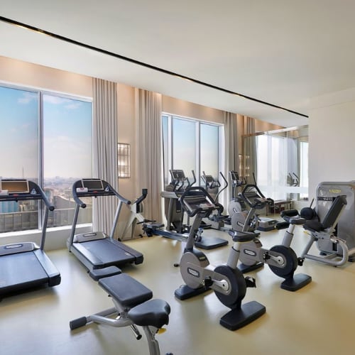 Ein modernes Fitnessstudio mit Kardio-Geräten, darunter Laufbänder, ein stationäres Fahrrad und Crosstrainer, in einem gut beleuchteten Raum mit großen Fenstern, die einen Blick auf die Stadt zeigen.
