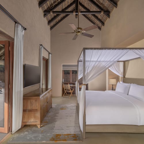 Luxuriöses Schlafzimmer in einer Villa mit Himmelbett, das mit weißen transparenten Vorhängen drapiert ist, einem großen an der Wand montierten Fernseher und einer offenen Tür, die zu einem Außenpool mit Liegestühlen führt.