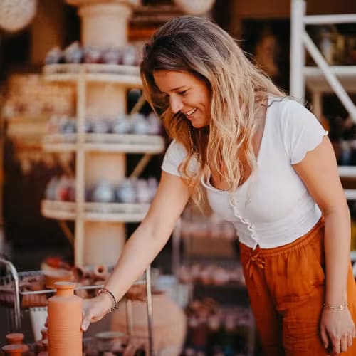 A woman shopping for pottery in a craft store, smiling and reaching for an item on a display table.