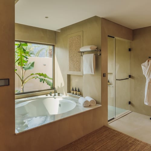 Modernes Badezimmer mit großer eingelassener Badewanne gefüllt mit Wasser, aufgerollten Handtüchern am Rand, verglaster Dusche, zwei weißen Bademänteln an der Wand und Fenster mit Blick auf grünen Pflanzen.