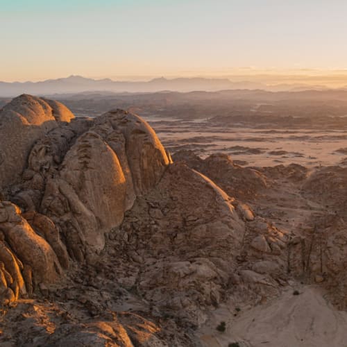 Sonnenuntergang über einer felsigen Wüstenlandschaft in Saudi-Arabien mit Bergen im Hintergrund.