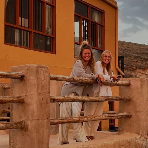 Two women standing on a balcony in front of an ochre-colored building, smiling at the camera during cloudy weather.