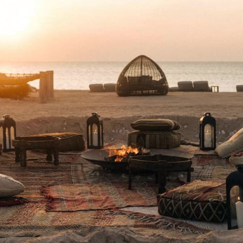 A cozy beach setting at sunset with cushioned seating around a fire pit, overlooking the sea with wicker lounge pods in the background.