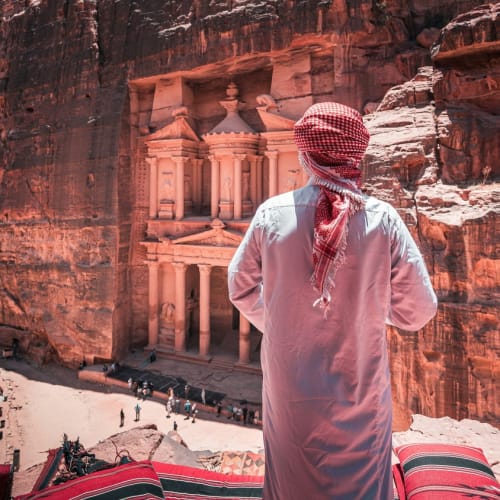 Ein Mann in traditioneller Kleidung des Nahen Ostens steht mit dem Rücken zum Kamera, blickt auf die antike Stadt Petra, die in rote Felswände geschnitzt ist.