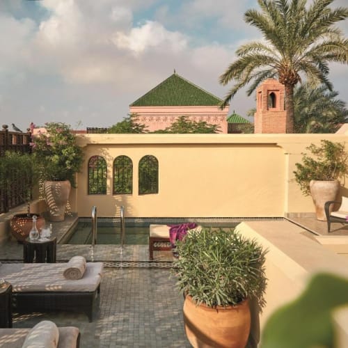 A luxurious rooftop terrace with lounge chairs, potted plants, and a small pool, overlooking a wall and traditional buildings with green rooftops and a palm tree.
