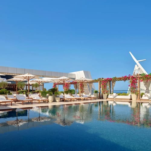 Luxurious outdoor pool area with sun loungers and umbrellas on a rooftop, overlooking the city skyline under a clear blue sky.