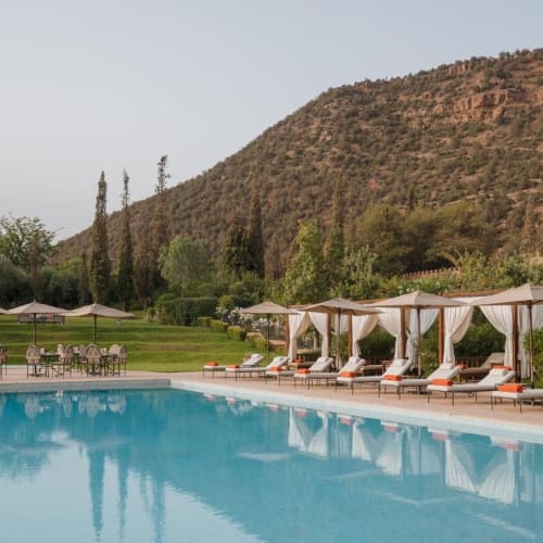 Entspannende Sonnenliegen mit weißen Vorhängen und einem Pool mit klarem Wasser, vor einer Kulisse aus grünen Bäumen und einem Berg.