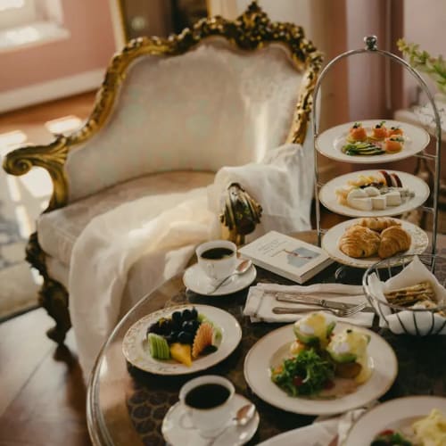 Ein luxuriöses Frühstück mit Kaffee, frischem Obst, Eggs Benedict, Croissants und verschiedenen Gerichten auf einem Glastisch neben einem verzierten, goldgerahmten Sessel mit weißer Decke in einem warm beleuchteten Zimmer.