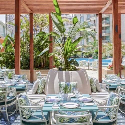  Elegant Außenessbereich des Four Seasons Hotel Cairo am Nile Plaza mit einem runden Tisch, der mit Geschirr, Glaswaren und bequemen Kissen geschmückt ist, umgeben von üppigem Grün und moderner Stadtansicht im Hintergrund.