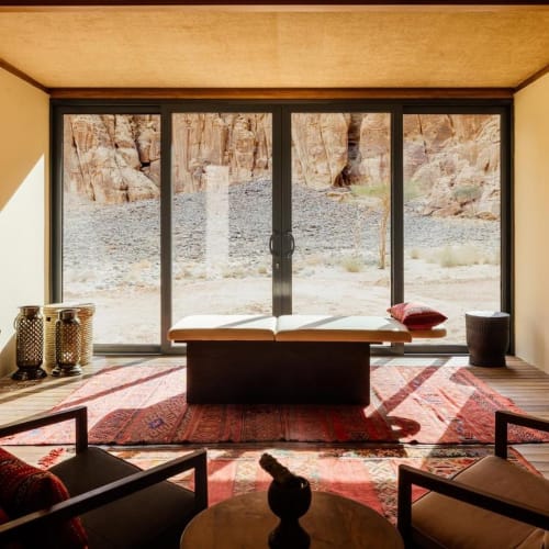 A serene spa treatment room with large floor-to-ceiling glass doors opening to a desert landscape outside, decorated with traditional rugs and modern furniture.