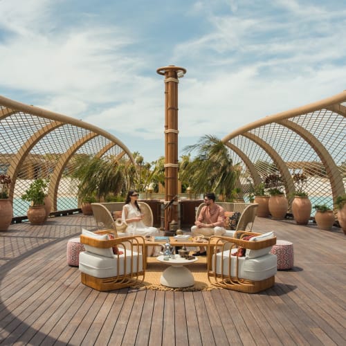Zwei Personen sitzen in einer kreisförmigen Anordnung von Stühlen und einem kleinen Tisch auf einer Dachterrasse mit moderner, halb offener Holzstruktur und Topfpflanzen unter einem bewölkten Himmel.