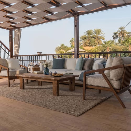 Outdoor lounge area with a wooden coffee table, cushioned sofa, and armchairs under a shaded pergola with tropical palm trees in the background.