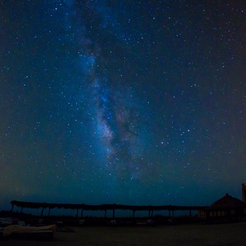 Night sky with a vivid view of the Milky Way galaxy above a dark, silhouetted eco-lodge structure.