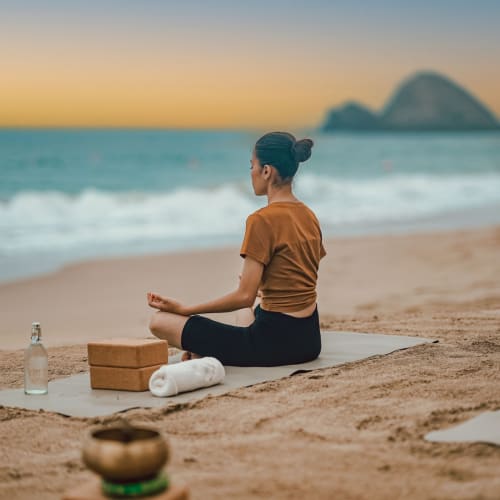 Eine Frau praktiziert Sitz-Yoga-Meditation auf einer Matte an einem Sandstrand bei Sonnenuntergang, mit einer Insel in der Ferne über dem Ozean.