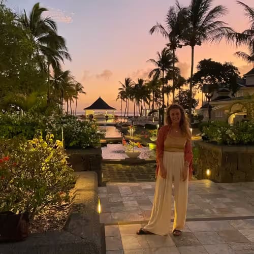 A woman stands on a stone pathway in a tropical garden at sunset, with palm trees, a gazebo, and the ocean in the background.