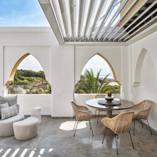 A modern terrace with a round table surrounded by woven chairs, a cushioned lounge chair with an ottoman, and arched windows offering views of palm trees and greenery under a partially covered roof.
