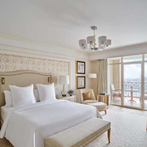 Luxurious hotel room with a large bed, bedside tables, lamps, and a seating area by the window with a balcony view of Cairo.
