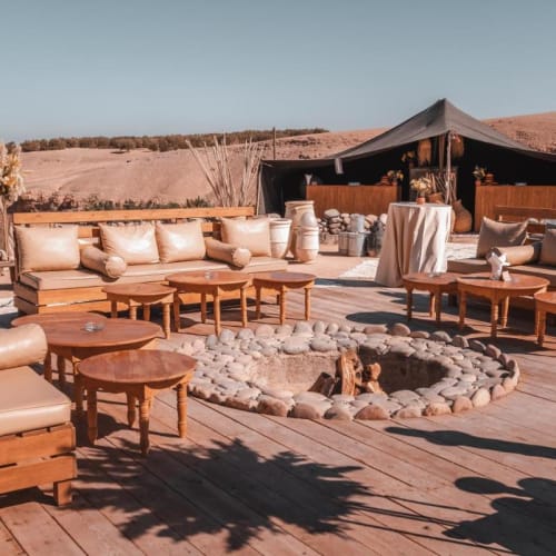 Lounge im Freien bei Inara Camp in Agafay mit Holzmöbeln um einen Feuerplatz, vor Wüstenlandschaft.