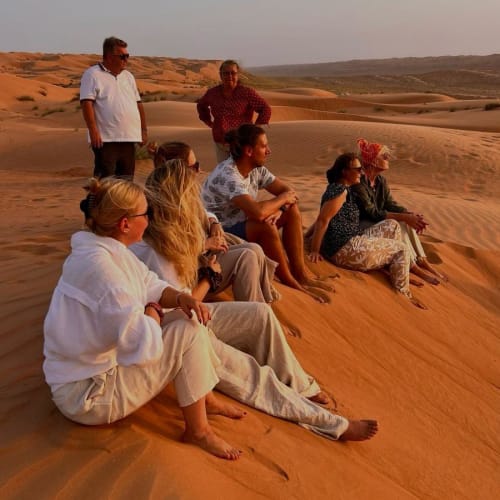 Group of people sitting on sand dunes in a desert during sunset.
