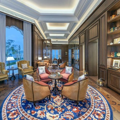 Elegante Lounge mit bequemen Stühlen, einem runden Tisch und einem großen dekorativen Teppich, in einem luxuriösen Hotelzimmer mit Bücherregalen und großen Fenstern.