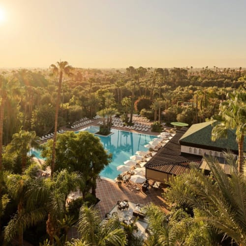 Luftaufnahme des La Mamounia Resorts mit Poolbereich, umgeben von üppigem Grün und hohen Palmen bei Sonnenuntergang.