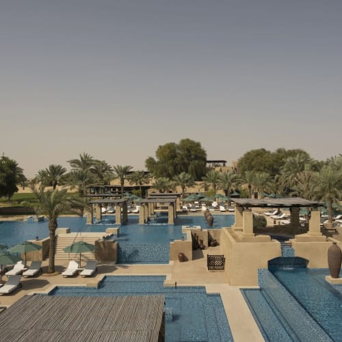 A luxurious desert resort with multiple pools, lounge chairs, and lush palm trees, under a clear sky.