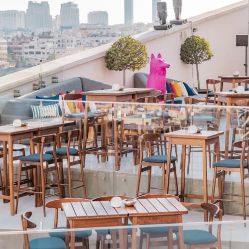 Eine Dachterrasse mit Holztischen und Stühlen, Dekokissen, Topfpflanzen und einer pinken aufblasbaren Tierfigur mit Blick auf eine Stadtskyline.