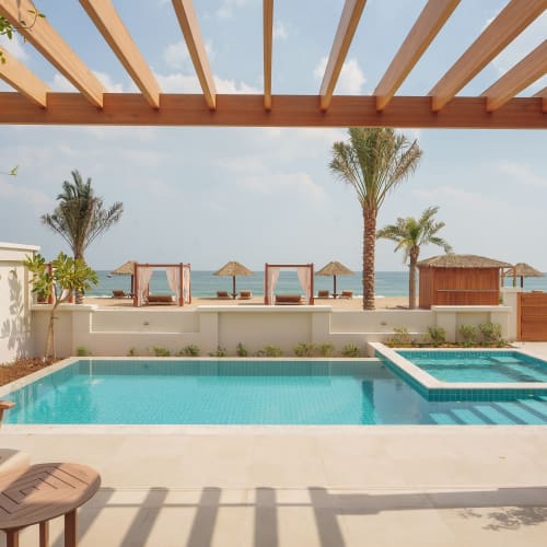 Ein ruhiger Außenbereich mit einem rechteckigen Schwimmbecken und angrenzendem Jacuzzi unter einer Holzpergola, mit Blick auf Palmen und Sonnenschirme am Strand.