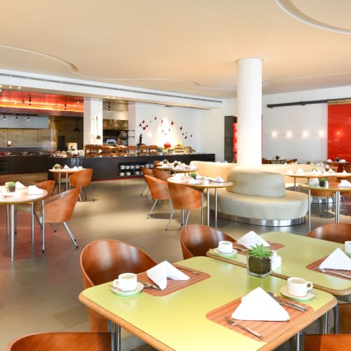 Elegant restaurant interior with round and square tables set for dining, featuring white cups, napkins, and small plants, with warm lighting and modern decor.