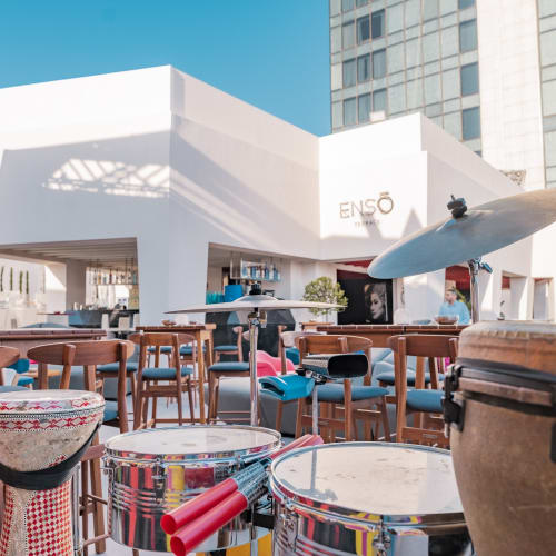 Außen-Terrasse im Enso-Restaurant mit bunten Trommeln, Stühlen und Sonnenschirmen in einer städtischen Umgebung.