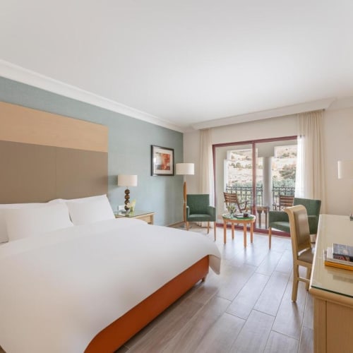 A spacious hotel room with a large bed, two nightstands, a sitting area with chairs and a balcony in the background.
