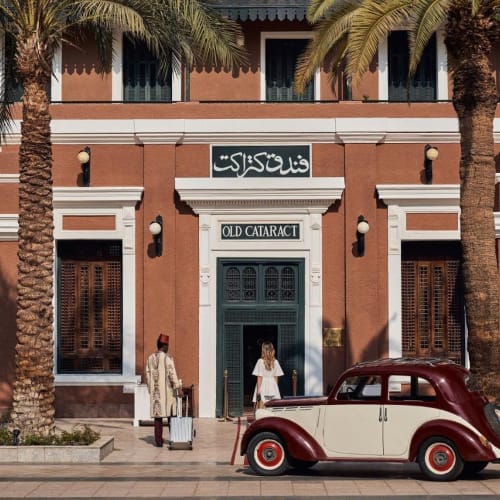 The entrance of the Old Cataract Hotel in Aswan, with palm trees, two people, and a vintage car in front.
